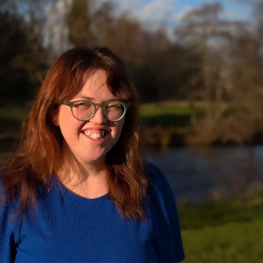 A person with long, reddish-brown hair and glasses smiles outdoors, wearing a bright blue shirt. The background features trees, grass, and a river under sunlight.