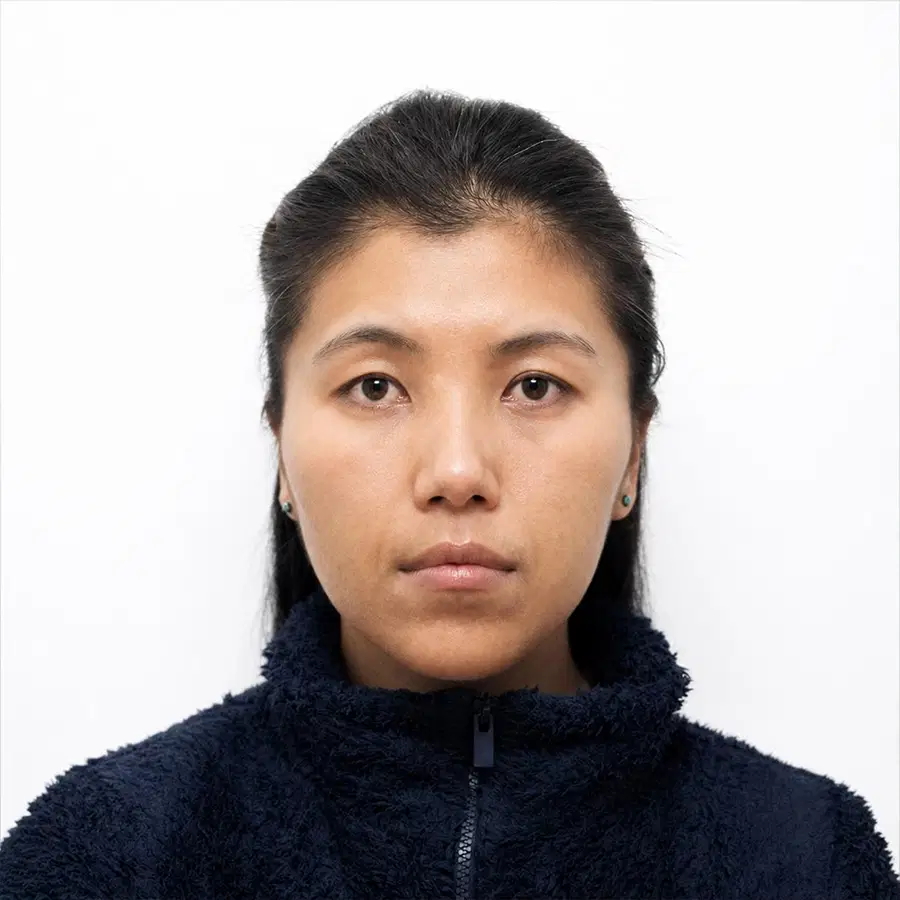 A person with long dark hair pulled back, wearing a dark, fuzzy zip-up jacket, looks directly at the camera with a neutral expression against a plain white background.