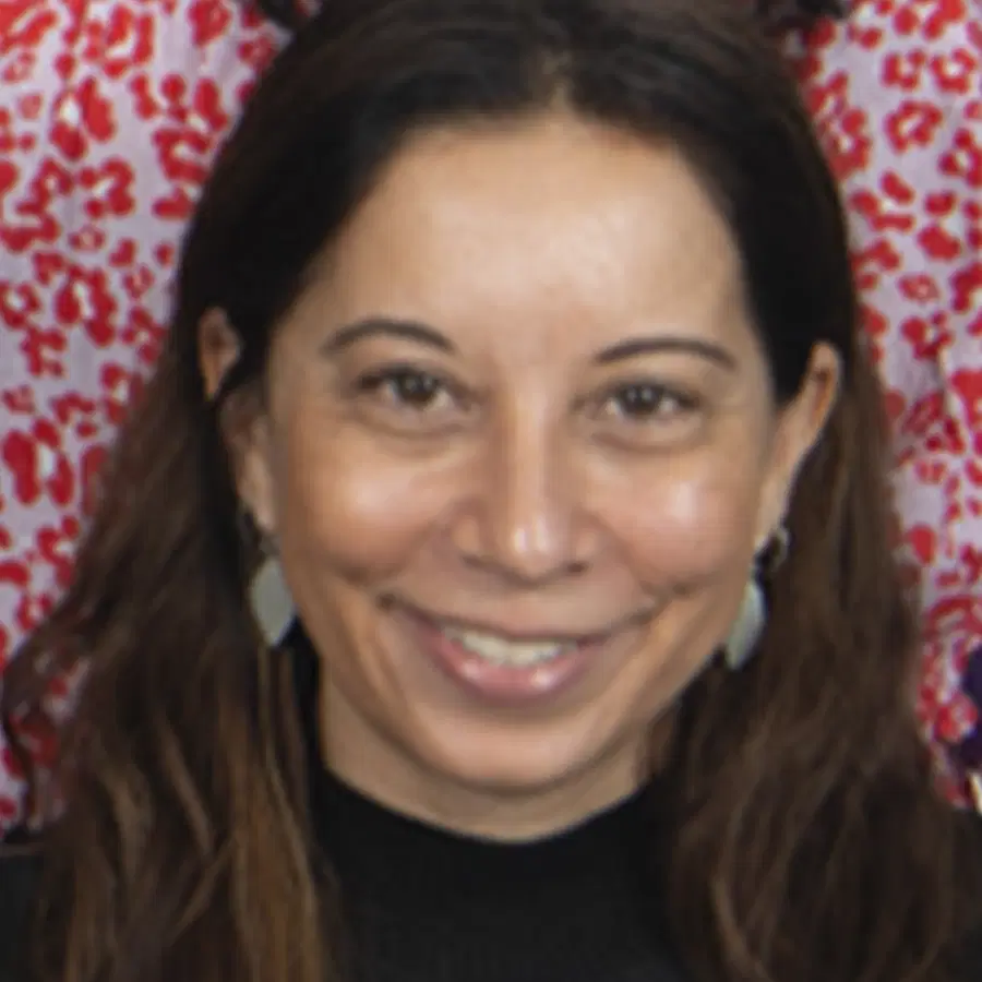 A woman with long brown hair smiles at the camera, wearing earrings and a black top. The background is red with white leopard-like spots.