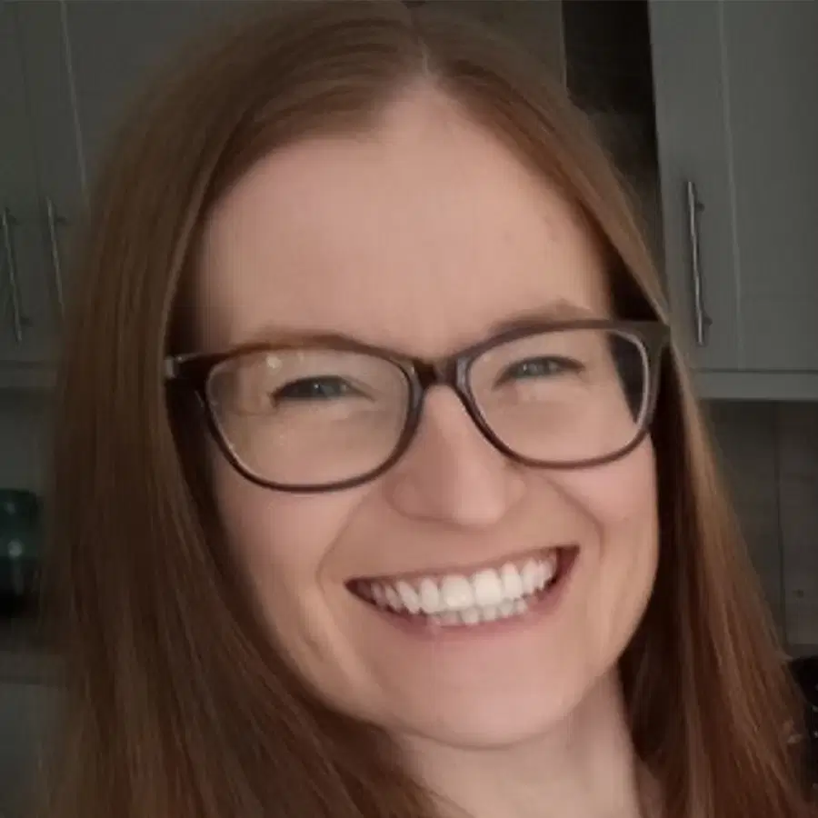 A person with long brown hair and glasses smiles widely, showing teeth. Kitchen cupboards and part of a worktop are visible in the blurred background.