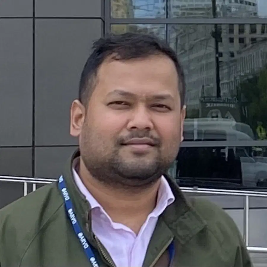A man with short dark hair and a beard, wearing a green jacket and white shirt, stands outdoors in front of a modern glass building.
