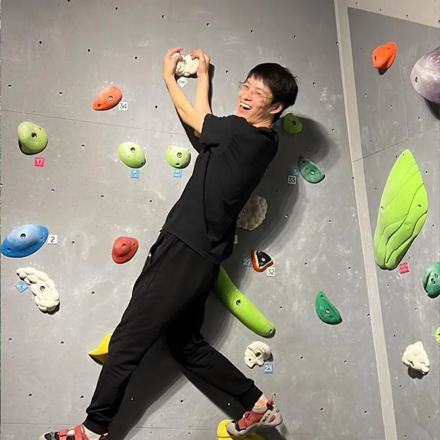 A person in black sportswear and climbing shoes smiles whilst hanging onto an indoor climbing wall with colourful holds.