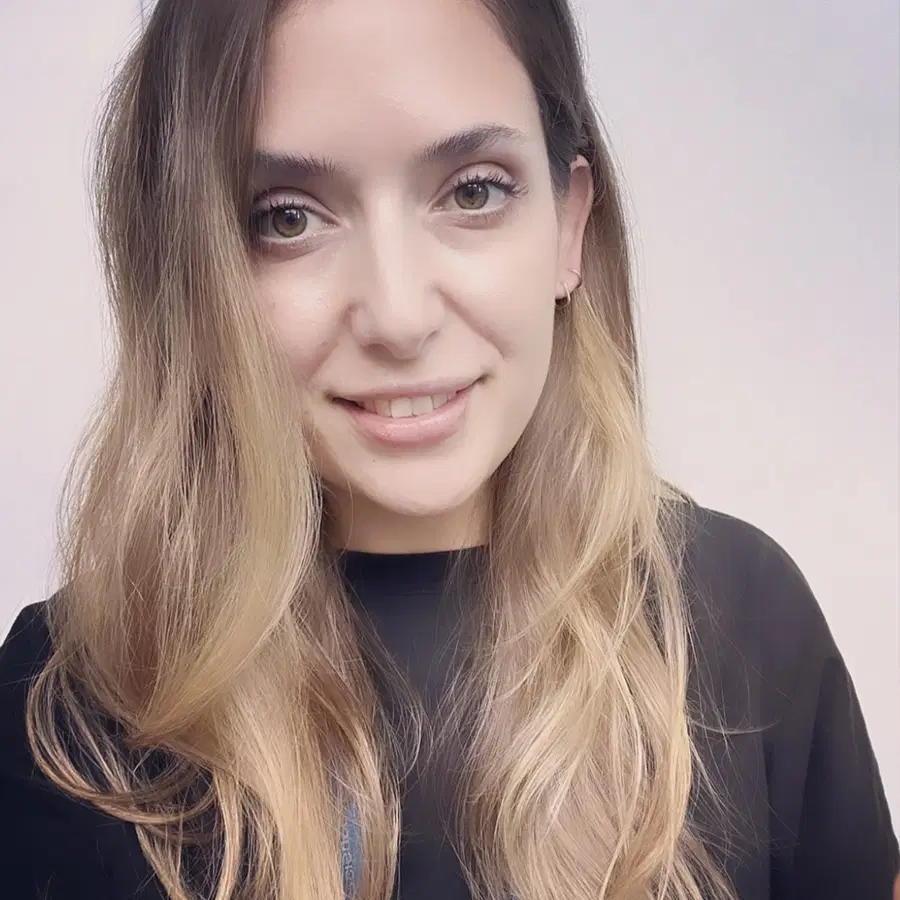 A woman with long, wavy, light brown hair and wearing a black top smiles softly at the camera against a plain, light background.