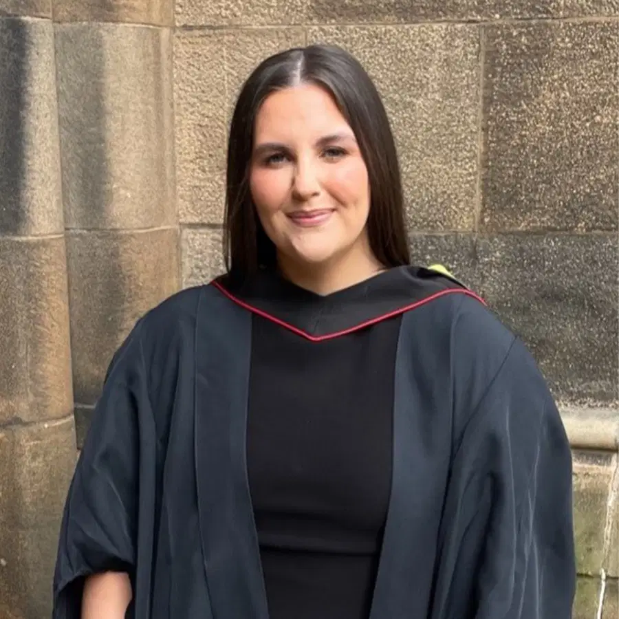 A woman in a black dress and graduation gown stands smiling in front of a stone wall.