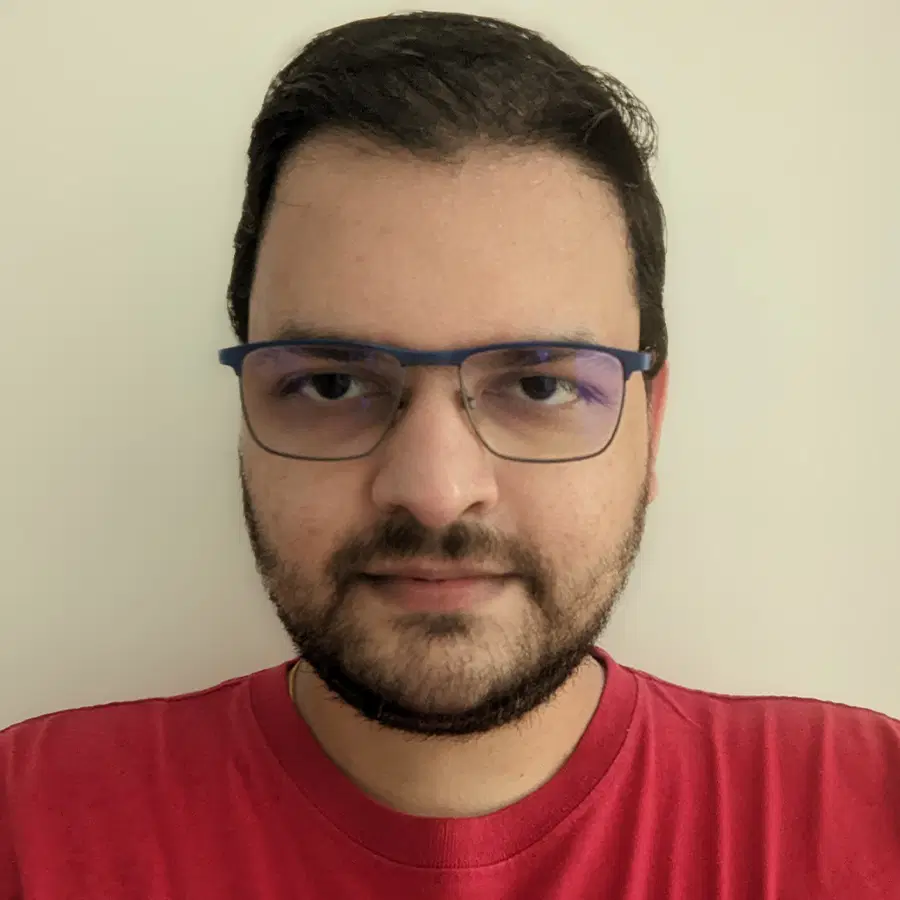 A man with short dark hair, glasses, and a trimmed beard wearing a red shirt stands in front of a plain light-coloured background, looking at the camera.