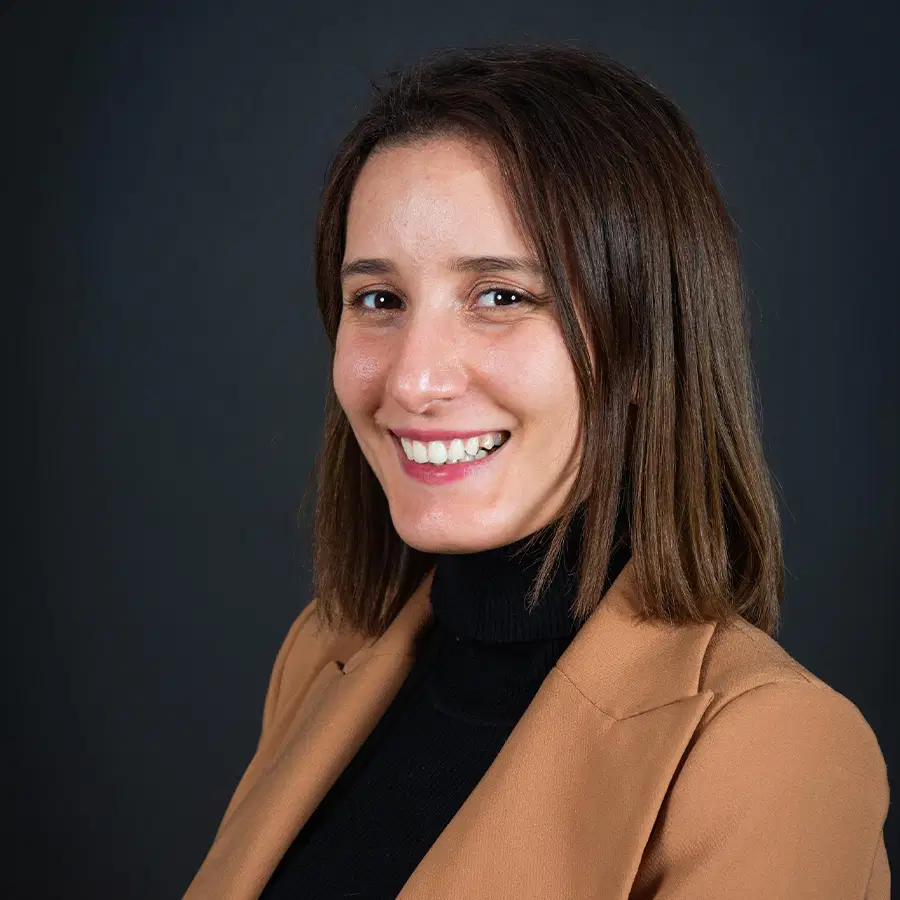 A woman with straight brown hair smiles at the camera. She wears a tan blazer over a black polo neck. The background is dark and plain.
