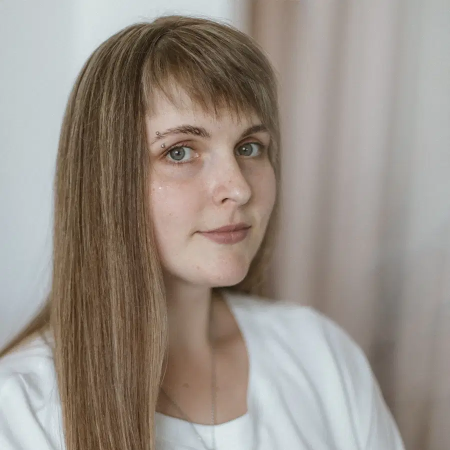 A woman with long straight brown hair, wearing a white shirt, looks at the camera with a neutral expression. She has an eyebrow piercing and is standing indoors with a blurred light background.