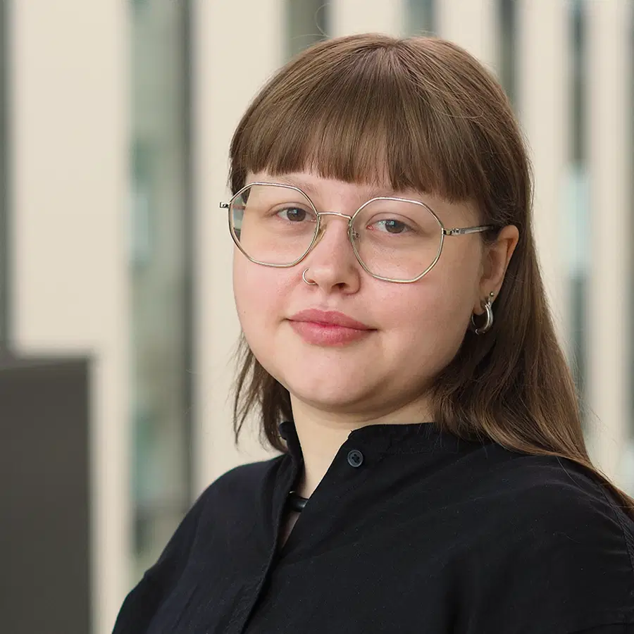 A person with straight brown hair, glasses, and a nose ring is wearing a dark top and looking at the camera. The background is softly blurred with vertical light and dark panels.