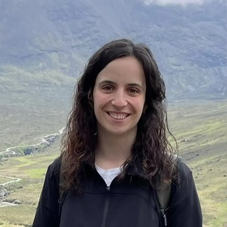 A woman with long, wavy brown hair smiles outdoors. She wears a black jacket and stands in front of a hilly, mountainous landscape under cloudy skies.