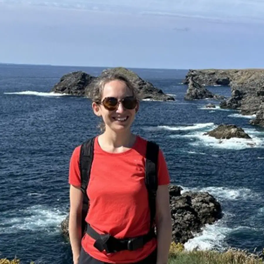 A person wearing sunglasses and a red shirt stands smiling near rocky cliffs and blue ocean water, with waves and a partly cloudy sky in the background.