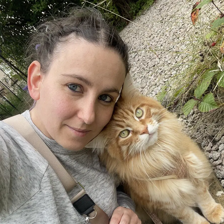 A woman with dark hair in a plait and a grey sweatshirt takes a close selfie outdoors with a fluffy ginger and white cat. Gravel and green plants are visible in the background.