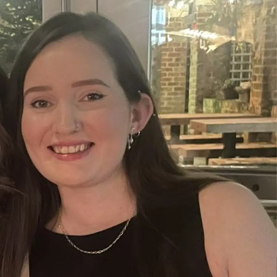 A young woman with long dark hair, wearing a sleeveless black top, a necklace, and earrings, smiles indoors with brick and wood décor visible in the background.