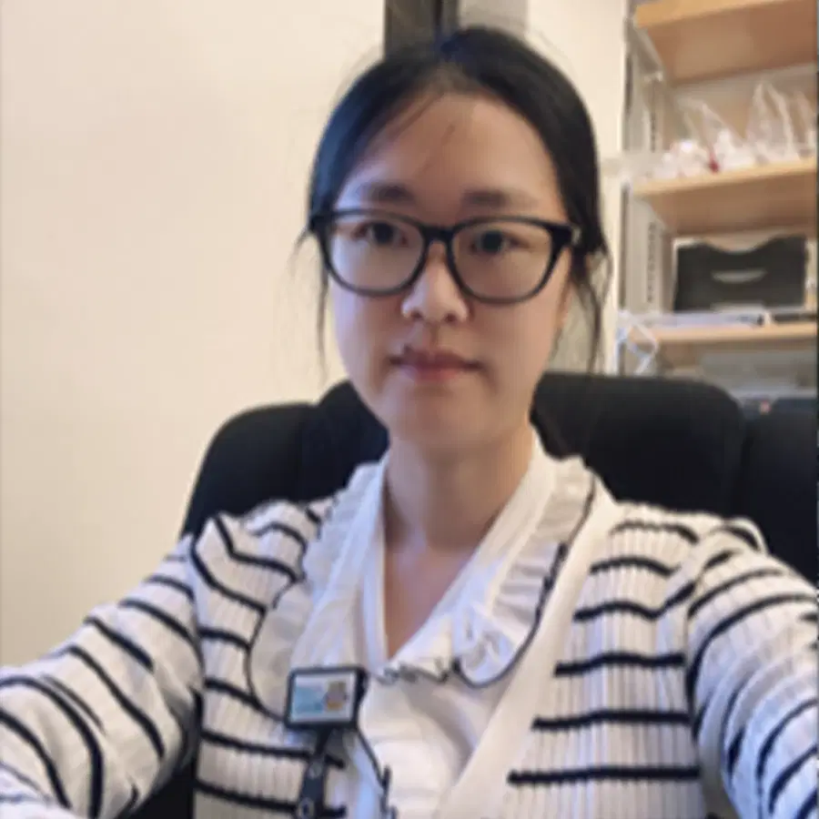 A person wearing glasses and a striped jumper sits in an office chair, looking at the camera. Shelves with various items are visible in the background.
