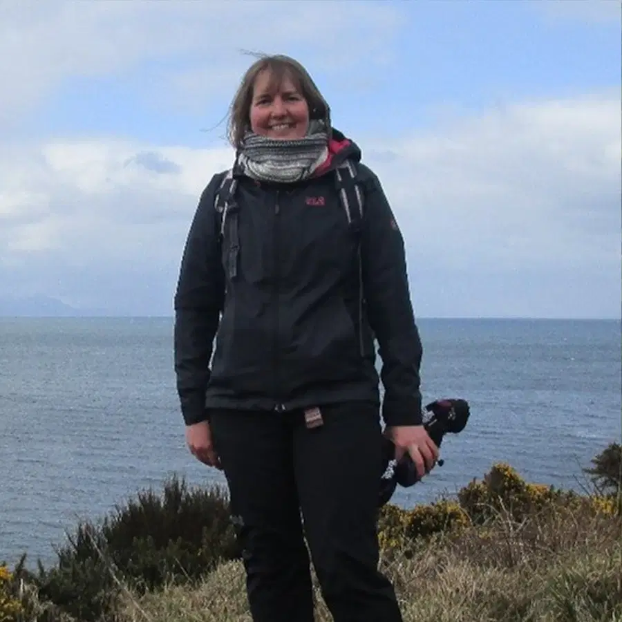 A person in a dark jacket and scarf stands outdoors near bushes, with the sea and a cloudy sky in the background.