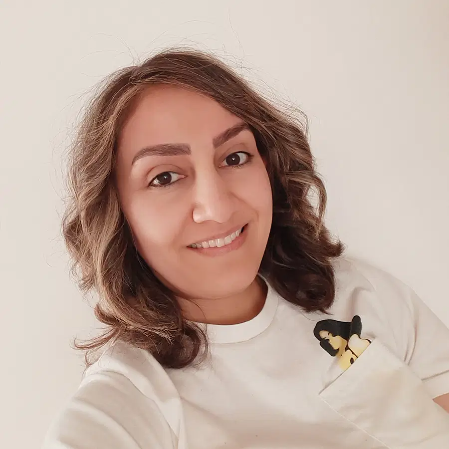 Woman with wavy brown hair smiling, wearing a white shirt with a cartoon woman design peeking out of the chest pocket, against a plain light background.