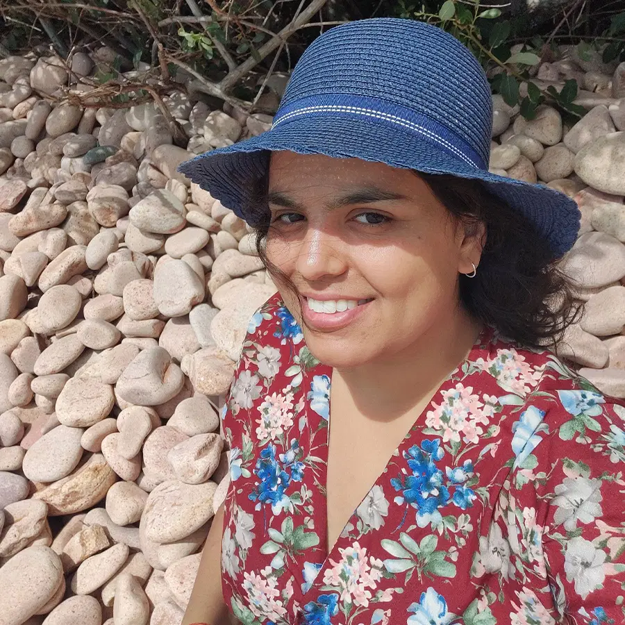 A person wearing a blue hat and floral dress smiles whilst sitting on large light-coloured pebbles outdoors, with green foliage visible in the background.
