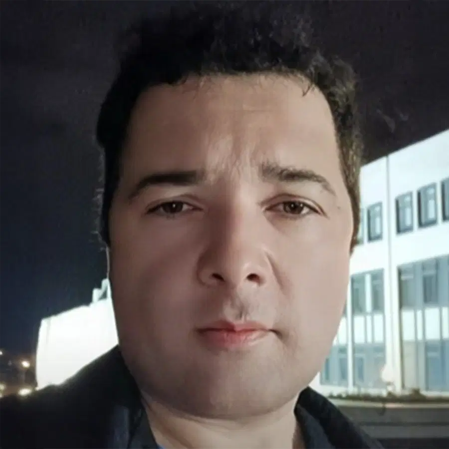 A person with short dark hair is standing outside at night in front of a modern, white building with large windows, looking directly at the camera.