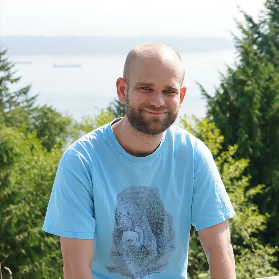 A smiling man with a beard and shaven head wears a blue T-shirt, standing outdoors with trees and a body of water in the background on a sunny day.
