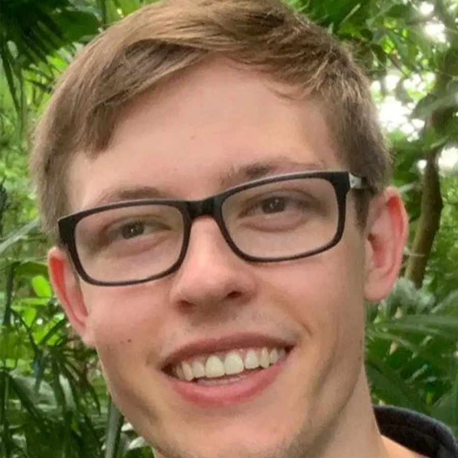 A person with short light brown hair and glasses is smiling outdoors, surrounded by green leafy plants.