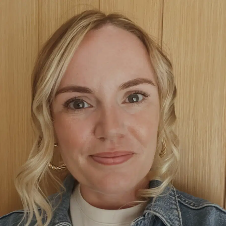A woman with wavy blonde hair, light skin, and blue eyes smiles softly. She is wearing a denim jacket and hoop earrings, standing in front of a wooden background.