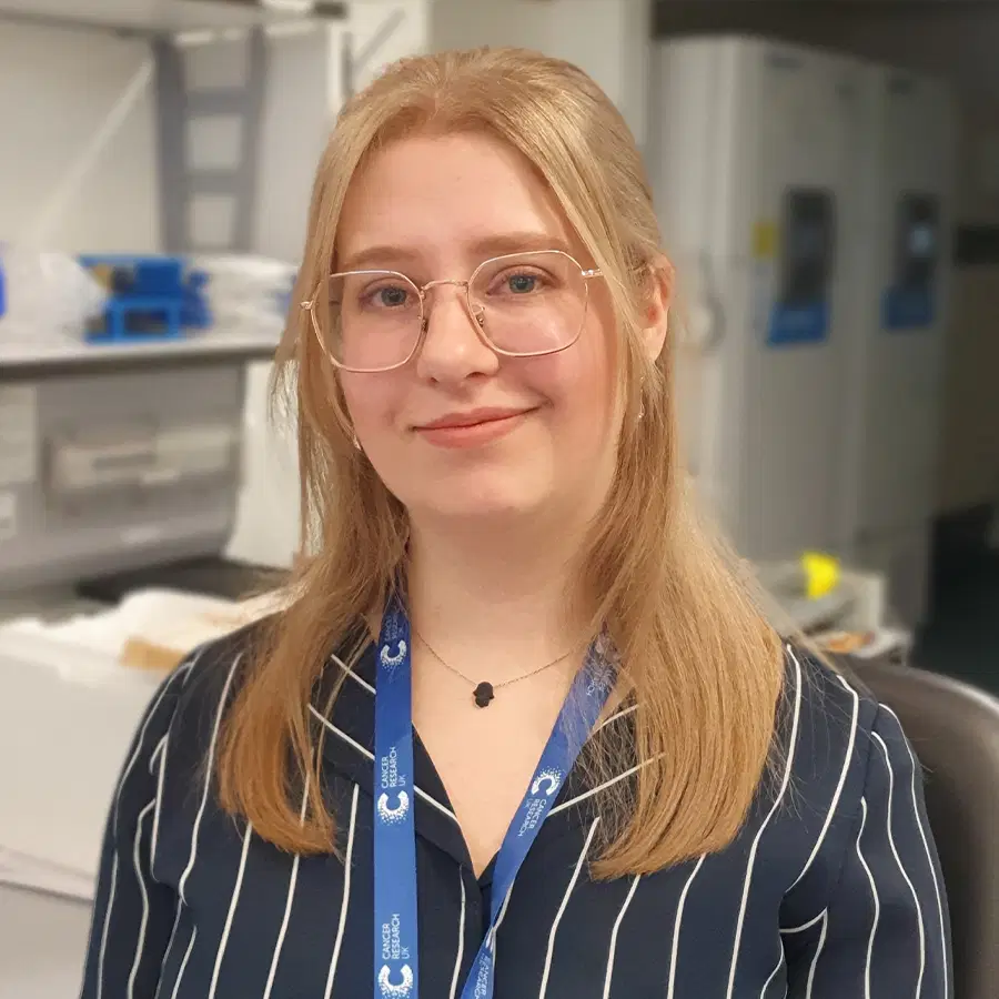 A person with fair hair and glasses, wearing a dark striped shirt and a blue lanyard, sits in an office or lab setting with shelves and equipment in the background.