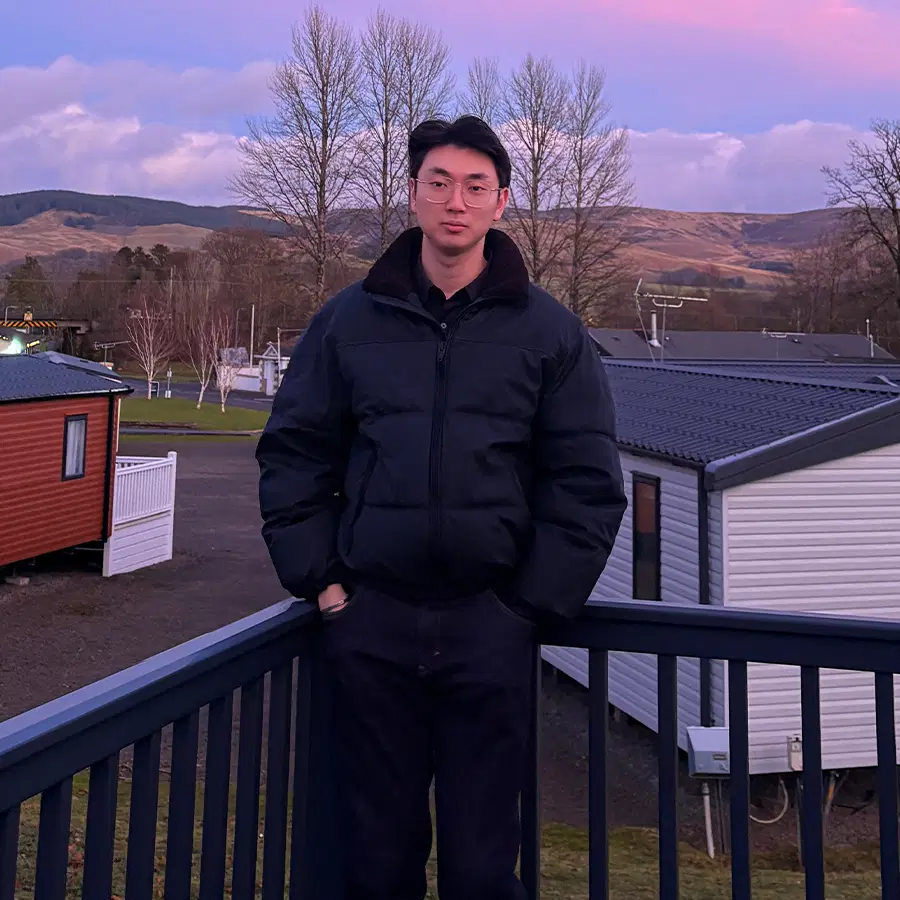 A person in glasses and a black jacket stands on a balcony with hands in pockets. Behind them are caravans, leafless trees, and hills under a pink and purple sky at sunset.
