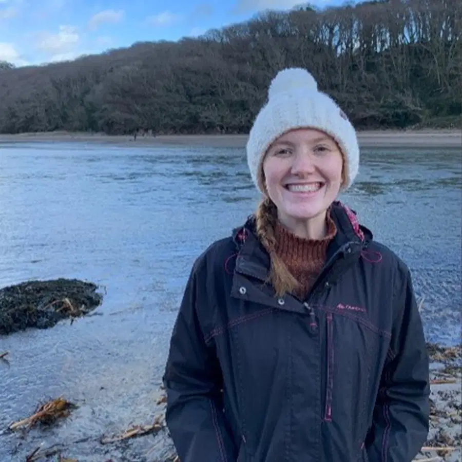 A smiling person in a dark jacket and white knitted hat stands by a river or lake, with a wooded hillside and cloudy blue sky in the background.