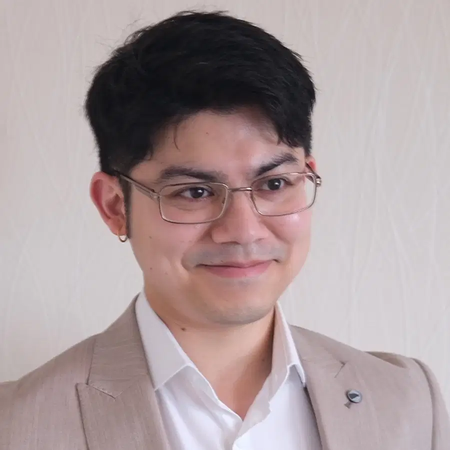 A man with short dark hair and glasses, wearing a light beige suit and white shirt, stands in front of a light patterned background, smiling slightly.