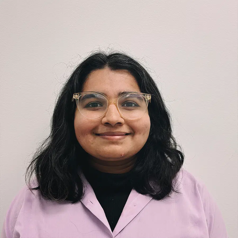 Person with shoulder-length dark hair wearing glasses and a lavender lab coat, standing against a plain light-coloured background, smiling slightly at the camera.