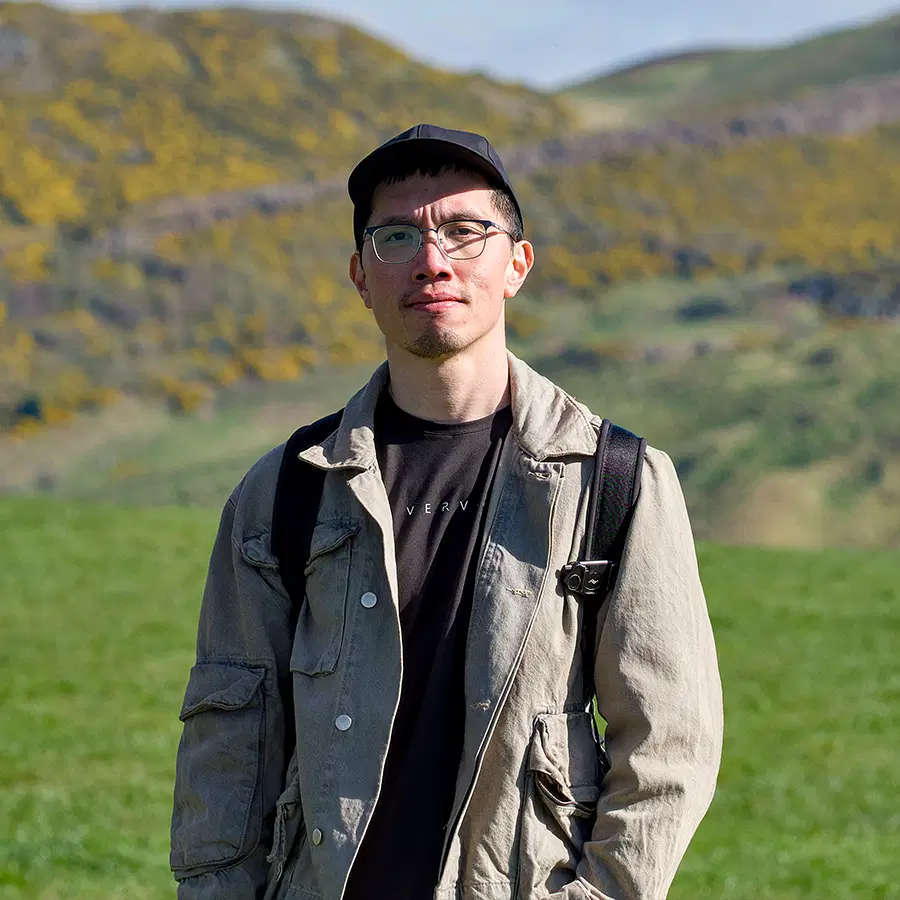 A person wearing glasses, a cap, and a light jacket stands outside on green grass with hills and flowers in the background, looking at the camera.