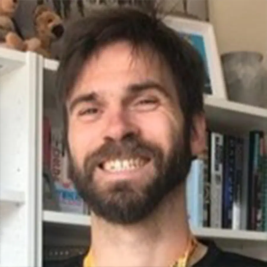 A bearded man with short hair smiles in front of a bookcase filled with books and a soft toy on the top shelf.