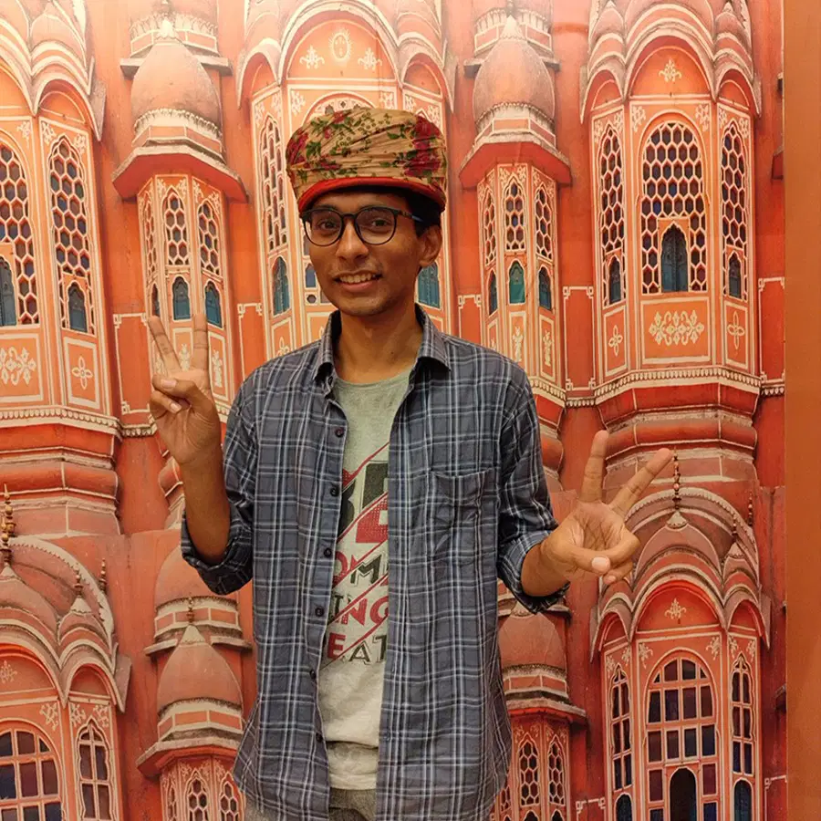 A person wearing glasses and a colourful turban makes peace signs with both hands, standing in front of an ornate red and pink architectural backdrop with arched windows.