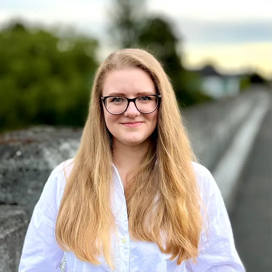 A young woman with long blonde hair and glasses stands outdoors, wearing a white shirt and smiling. A blurred background shows trees and a stone wall.