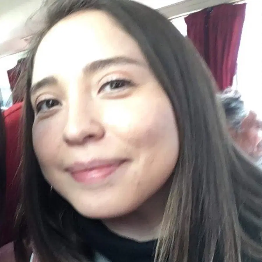 A person with long brown hair smiles slightly, looking at the camera. Red curtains are visible in the background.