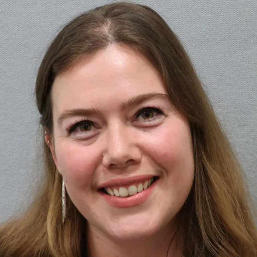 Woman with long brown hair smiling at the camera, wearing a light top and dangling earrings, against a plain light grey background.