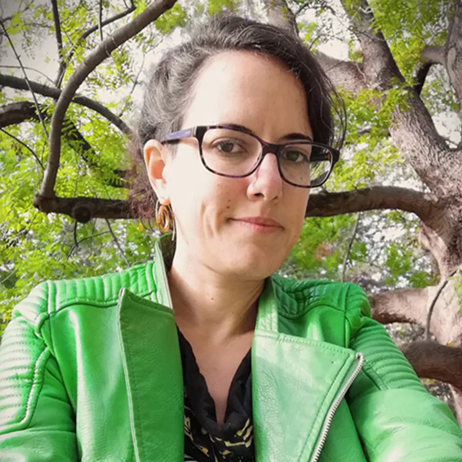 A person with glasses and brown hair wears a bright green jacket and black shirt, standing outdoors in front of leafy tree branches.