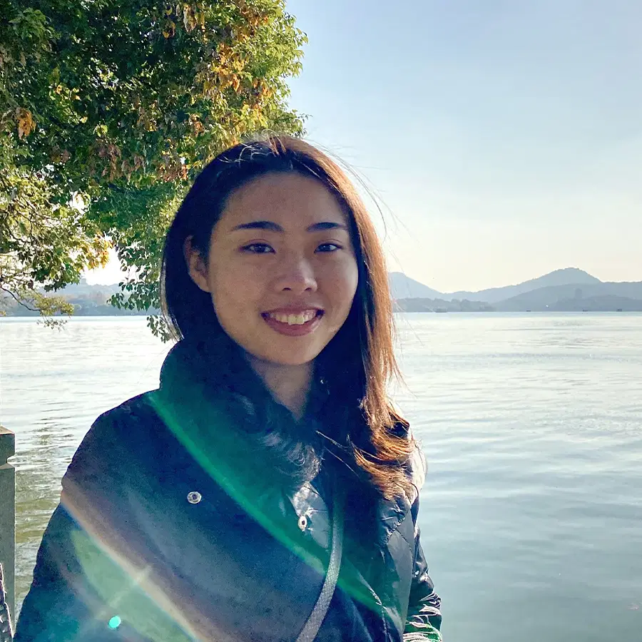 A woman smiles outdoors by a calm lake, with tree branches overhead, mountains in the distance, and sunlight creating a soft lens flare across the image.