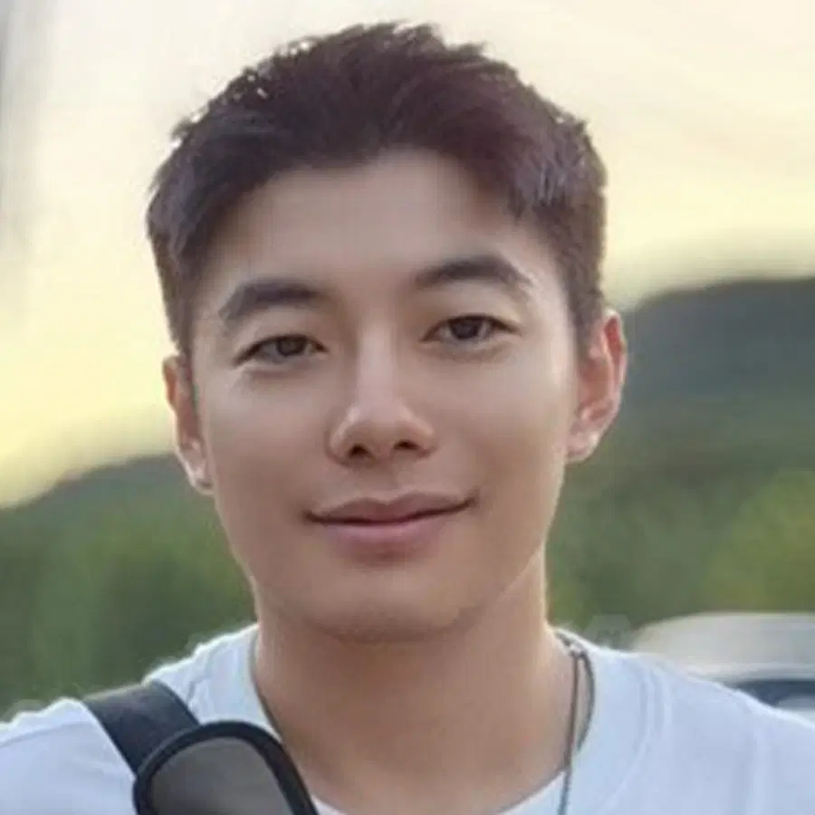 A young man with short dark hair smiles softly outdoors, wearing a white shirt and carrying sunglasses on his shoulder.