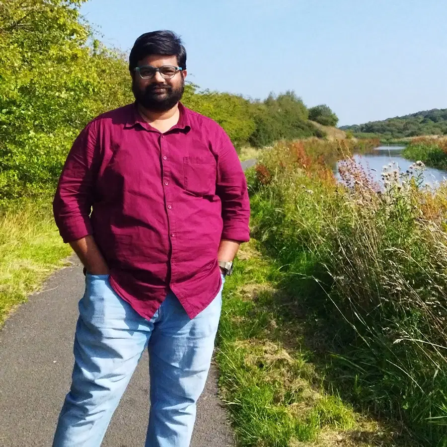 A man in a maroon shirt and light blue jeans stands on a path beside tall grass and a river, with trees and blue sky in the background.