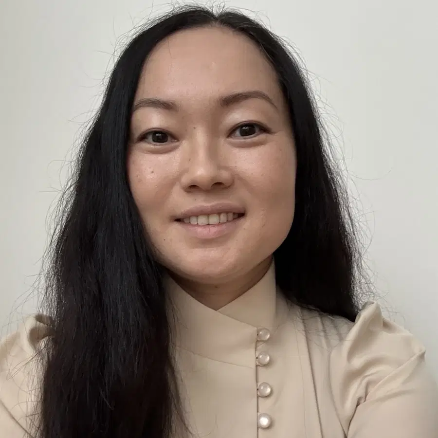 A person with long dark hair and light skin smiles at the camera, wearing a beige top with pearl buttons, against a plain light background.