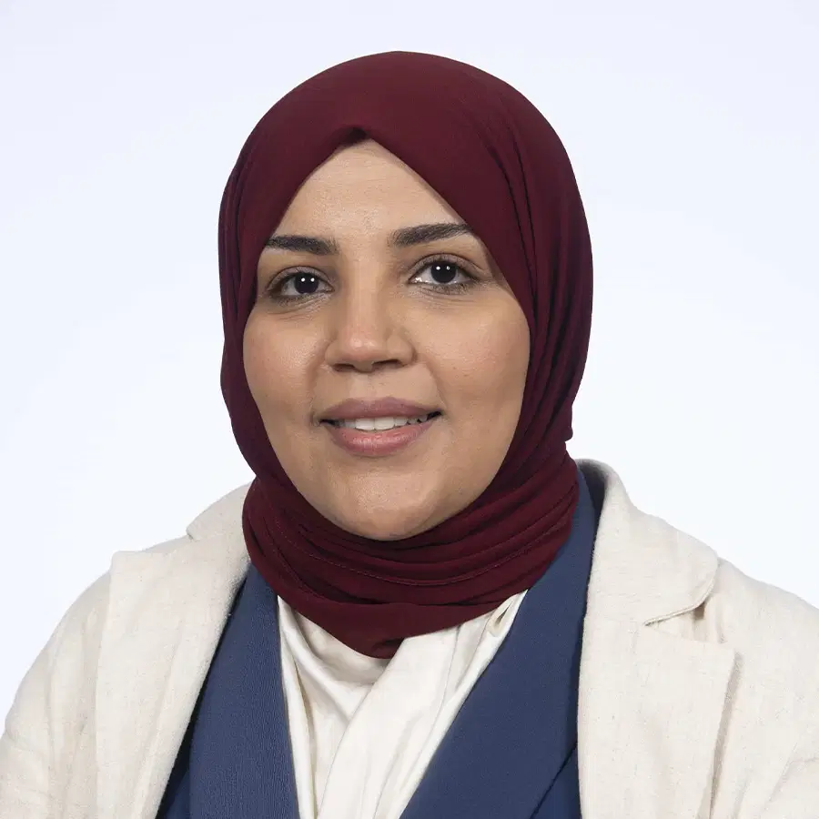 A woman wearing a maroon hijab, beige blazer, and blue waistcoat smiles at the camera against a plain light background.