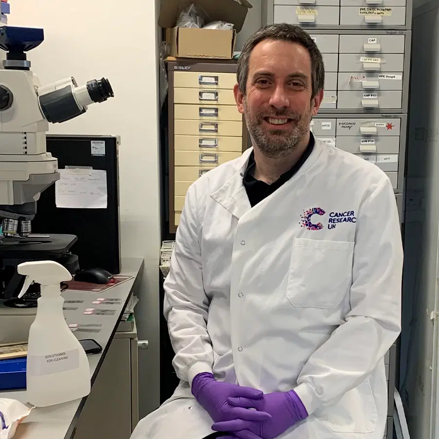 A person in a white Cancer Research lab coat and purple gloves sits in a laboratory beside a large microscope, with drawers and lab equipment visible in the background.