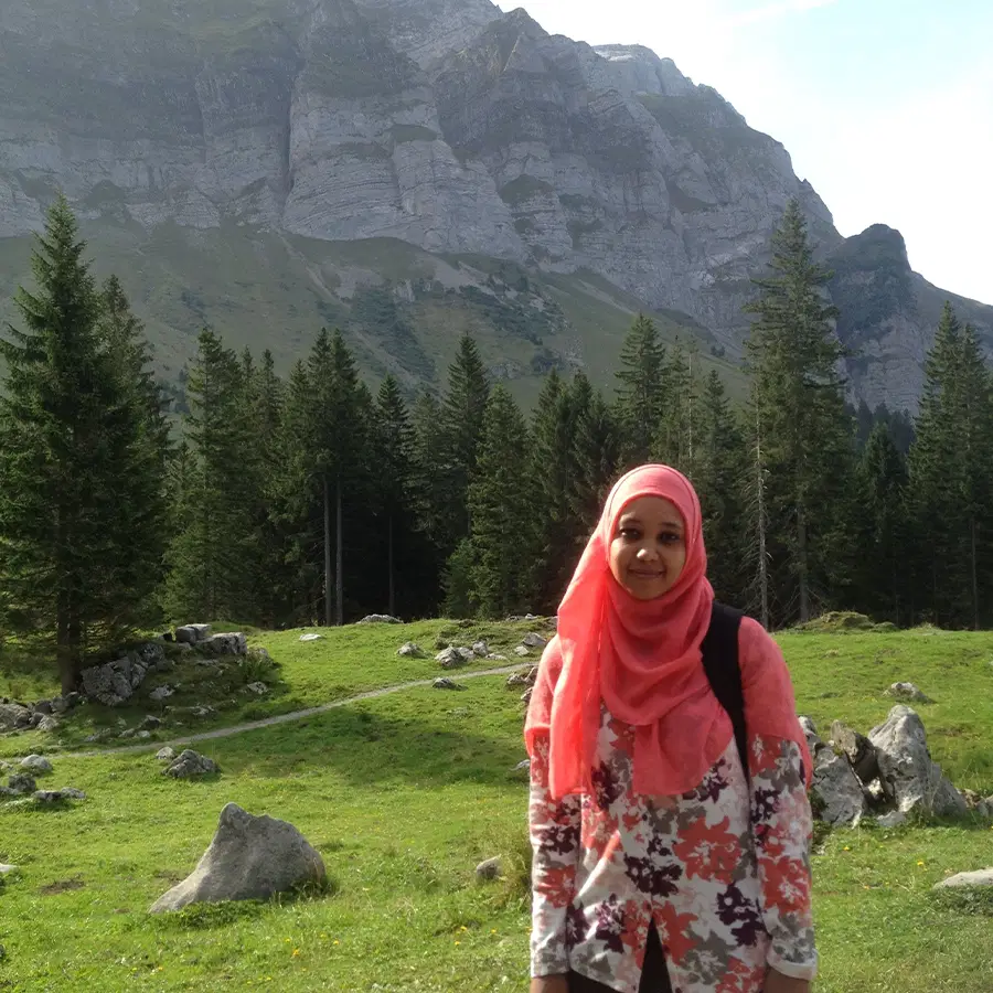 A woman in a pink hijab and floral jacket stands on a grassy field with rocks, pine trees, and tall mountains in the background, under a partly cloudy sky.