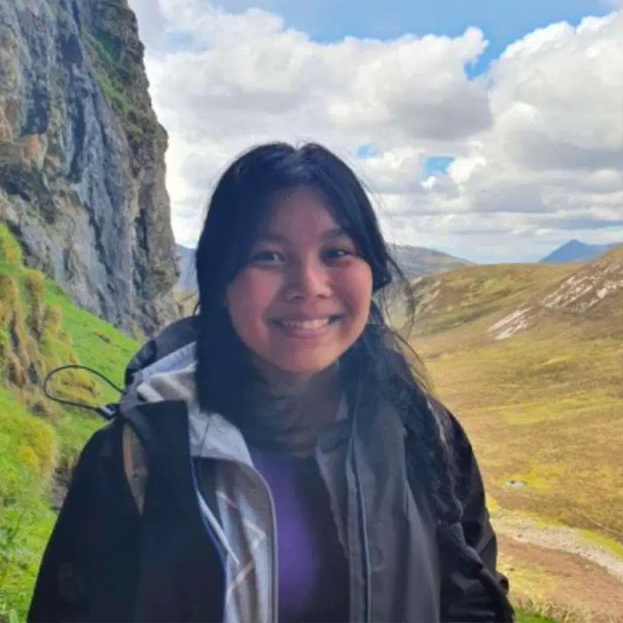 Young person with dark hair smiles at the camera, standing outdoors near a rocky cliff and grassy hills under a partly cloudy sky.