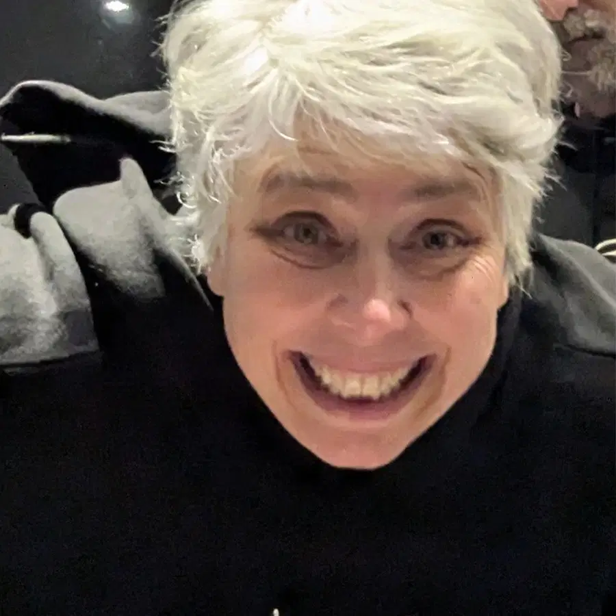 A person with short grey hair smiles widely at the camera, wearing a dark top, with another person partially visible in the background.