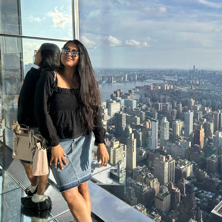 A woman in sunglasses poses by a glass wall with her reflection, overlooking a cityscape full of tall buildings on a sunny day.