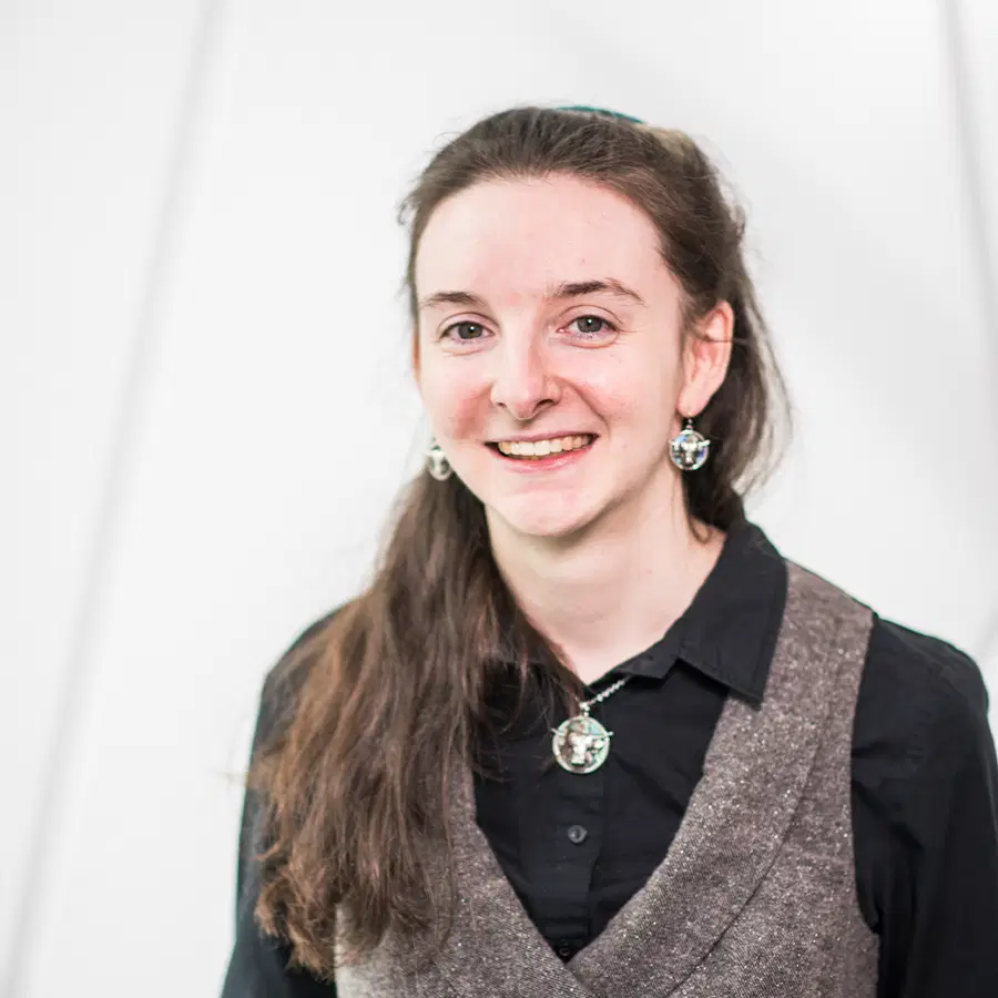 A person with long brown hair, wearing a black shirt, grey waistcoat, and matching earrings and necklace, smiles at the camera against a white background.