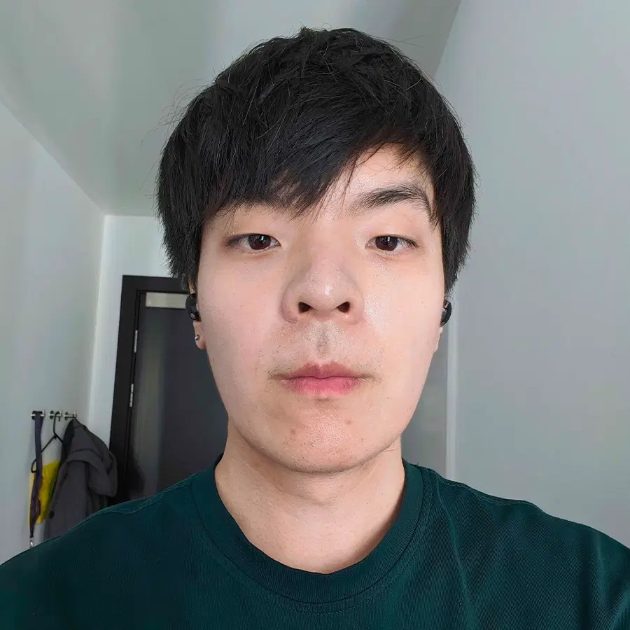 A person with short black hair wearing a dark green shirt looks directly at the camera, standing indoors near a door with coats hanging on pegs in the background.
