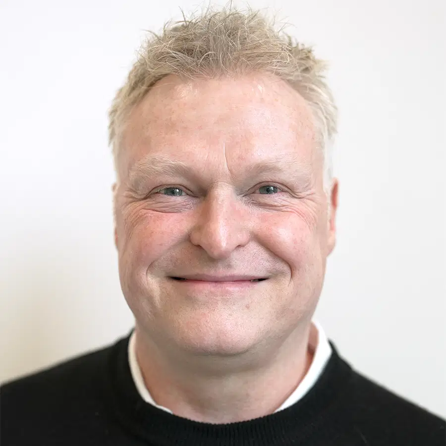 A middle-aged man with short, light-coloured hair smiles warmly at the camera. He is wearing a black jumper over a white collared shirt, and the background is plain and light-coloured.