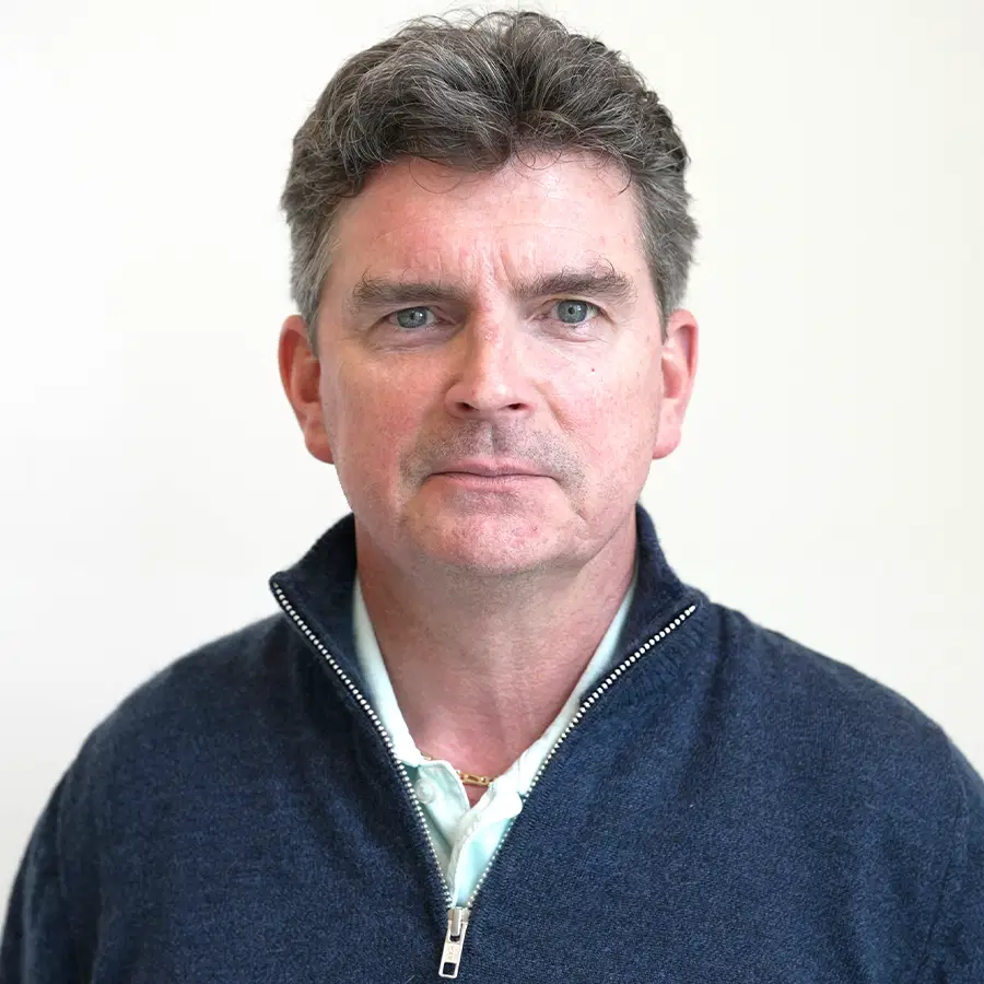A middle-aged man with short brown hair and blue eyes, wearing a blue zip-up jumper over a white collared shirt, stands against a plain white background.